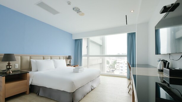 A hotel room with a large bed, a desk with a lamp, a TV, and a view of the city through large windows.
