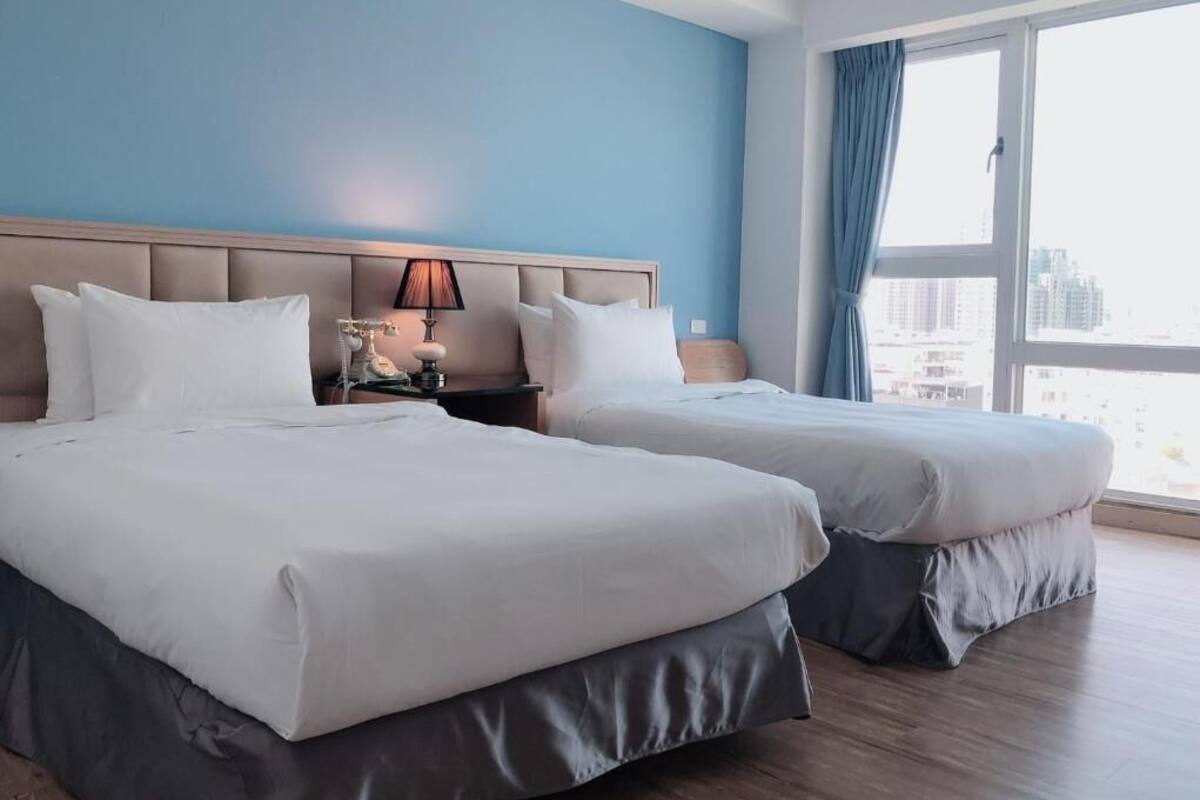 Una habitación de hotel con dos camas, una mesita de noche con lámpara y un ventanal con cortinas.