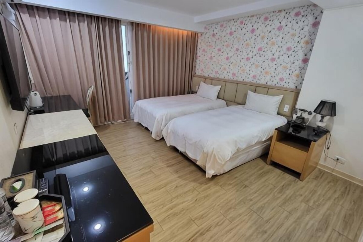 Habitación de hotel con dos camas, un escritorio, una silla, una lámpara y una mesita con una taza y una caja.