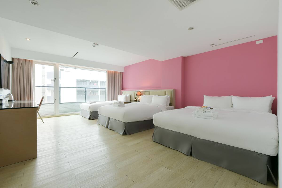Habitación de hotel con dos camas, un escritorio y vistas de la ciudad a través de amplios ventanales.