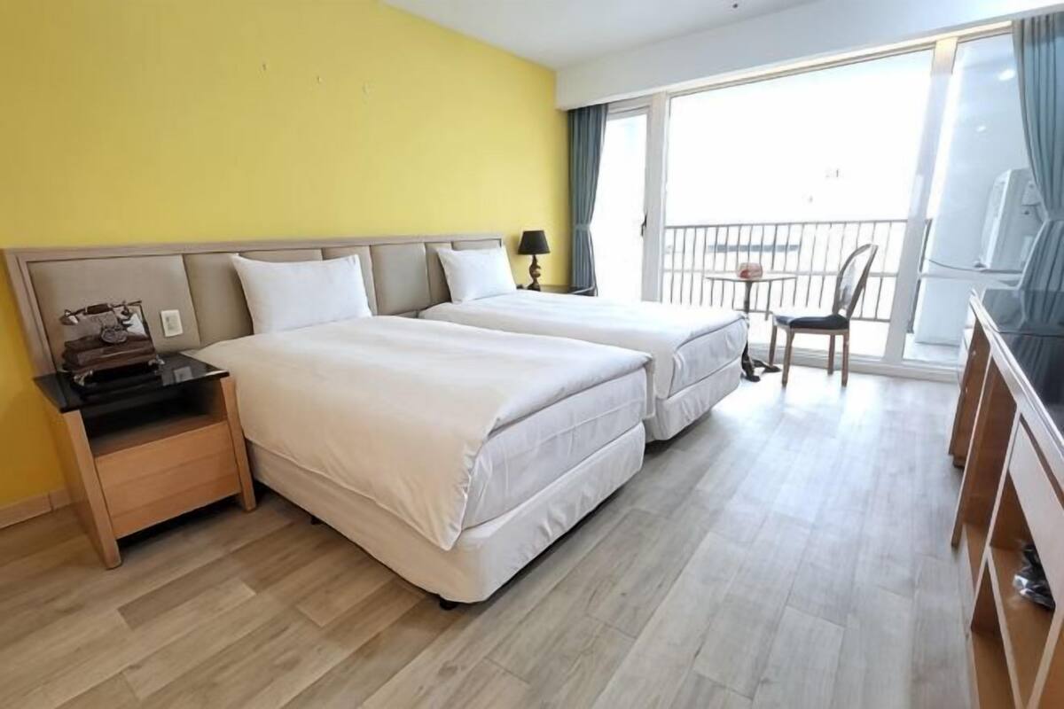 Habitación de hotel con dos camas, un escritorio, una silla y un balcón con barandilla.