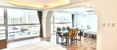 A hotel room with a large bed, a dining table with chairs, and a view of the cityscape through large windows.