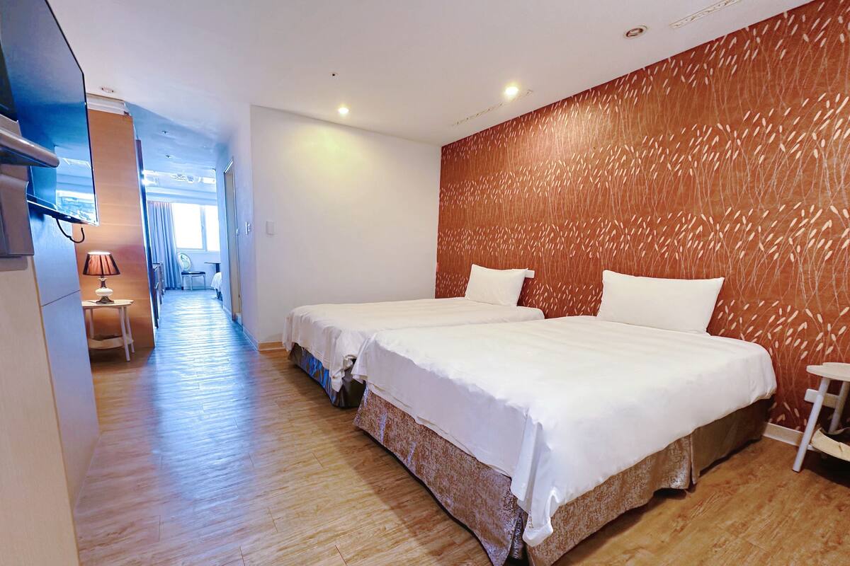 Habitación de hotel con dos camas, suelo de madera, pared con textura y una mesita con lámpara.