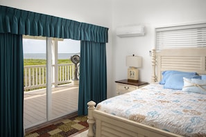 Un dormitorio con cama, mesita de noche, lámpara, ventana con persianas y vista al océano desde un balcón.