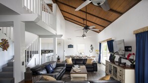 A spacious living room with a black leather sectional sofa, a wooden coffee table, and a ceiling fan. A staircase with white railings leads to an upper level. - Direct Ocean View! 5 Bed/3 Bath with room for everyone. Large pool and Dock! (Summerland)