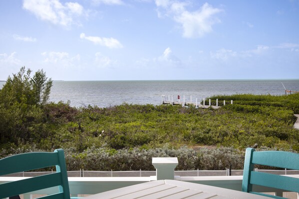 A view from a balcony overlooking the sea with a table and chairs, a railing, and some vegetation. - Direct Ocean View! 5 Bed/3 Bath with room for everyone. Large pool and Dock! (Summerland)