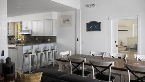 A modern kitchen with white cabinets, a dining area with a wooden table and white chairs, and a living room with a black sofa and a yellow pillow. - Direct Ocean View! 5 Bed/3 Bath with room for everyone. Large pool and Dock! (Summerland)