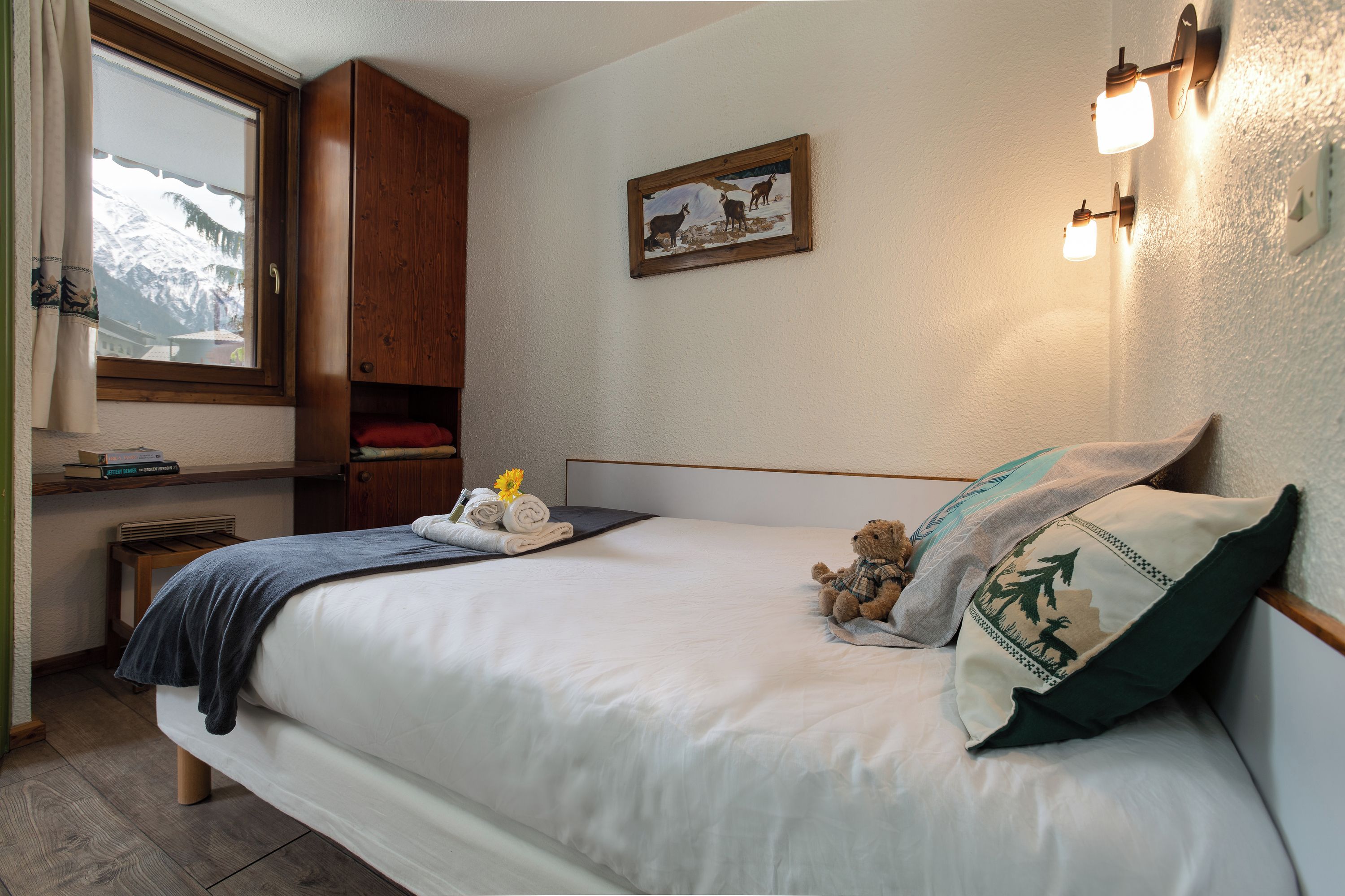A neatly made bed with a pillow and a teddy bear, a wooden wardrobe, a framed picture on the wall, and a window with a view of snow-capped mountains.