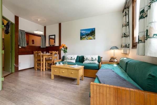 A cozy living room with a wooden floor, a green sofa, a wooden coffee table, and a dining area with a wooden table and chairs.