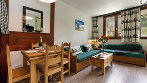 Een houten eettafel met stoelen, een groene bank, een bijzettafeltje met een bloevas en een raam met gordijnen.