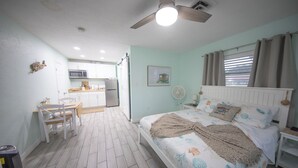 Ein Schlafzimmer mit Bett, Nachttisch und Ventilator. Es gibt eine Küchenzeile mit Schränken und einer Mikrowelle. Der Raum verfügt über einen Deckenventilator und ein Fenster mit Vorhängen.