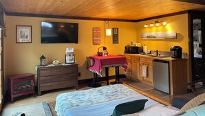 Een knus ingerichte kamer met een bed, een bureau met een rode tafelkleed, een kitchenette en een aan de muur bevestigde televisie.