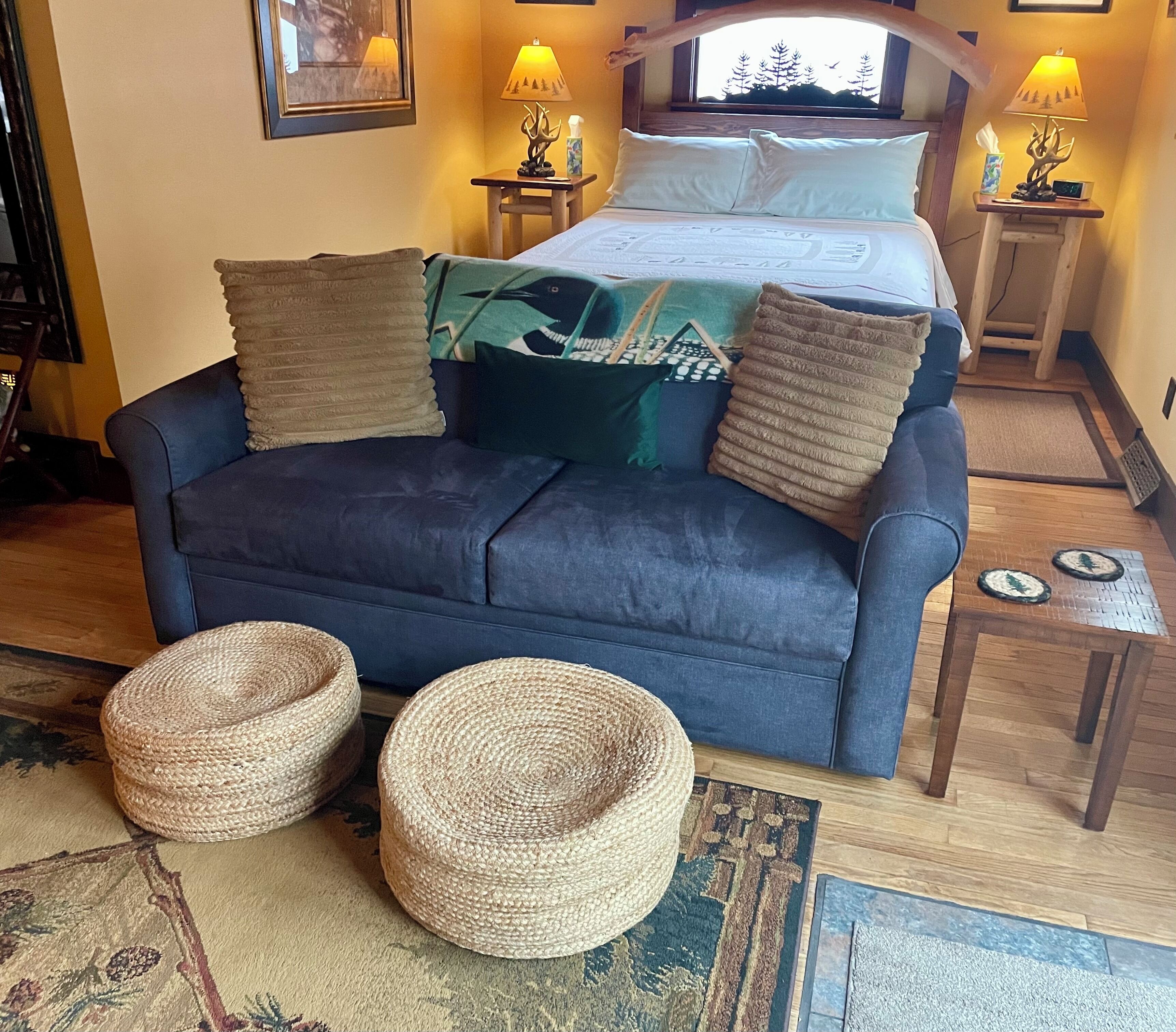 A cozy room with a bed, two nightstands, a blue sofa, two woven poufs, and a view of trees through the window.