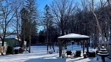Un kiosque agrémenté de bancs, le sol recouvert de neige, entouré d’arbres sans feuilles et surplombé d’un ciel dégagé.