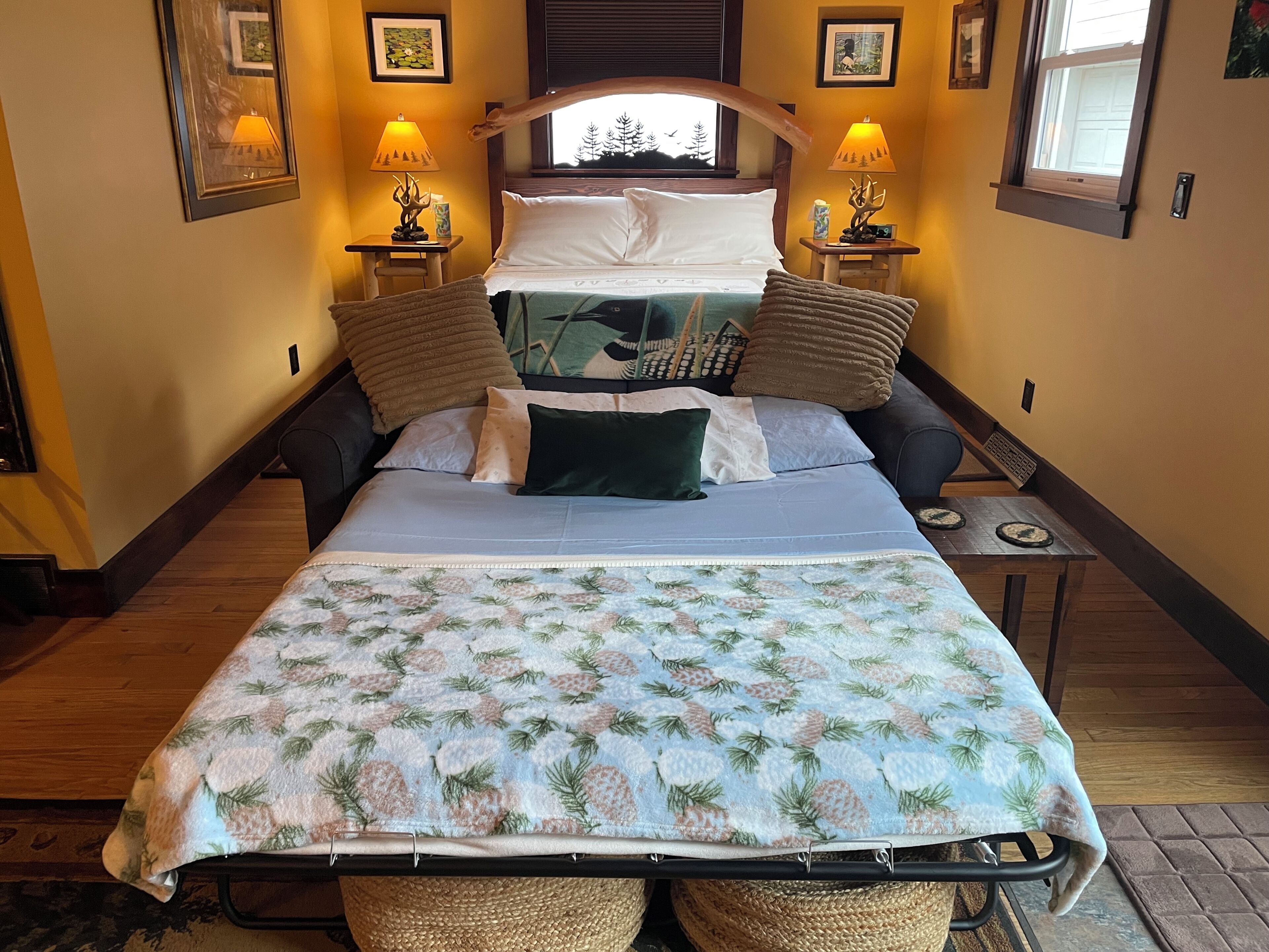 A bedroom with a bed, two bedside tables with lamps, a window with a view of trees, and a wall-mounted picture frame.