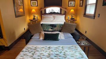 A bedroom with a bed, two bedside tables with lamps, a window with a view of trees, and a wall-mounted picture frame.