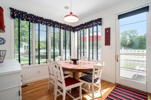 Un coin repas avec une table ronde, des chaises blanches, une nappe à rayures rouges et blanches, et une vue dégagée grâce à de grandes fenêtres équipées de volets.