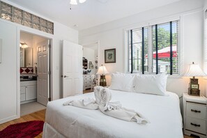 Une chambre d’hôtel avec un grand lit, une fenêtre avec des stores, une table de chevet avec une lampe et un tapis rouge.