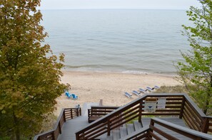 Un escalier en bois mène à une plage où se trouvent des chaises bleues et un panneau d’avertissement du service de surveillance des plages.