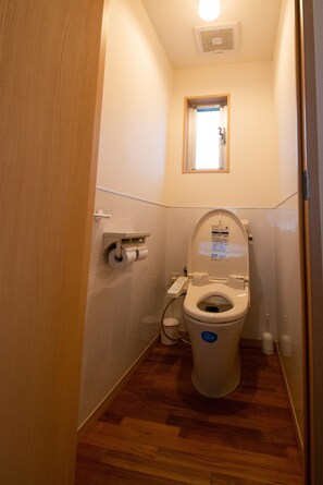 A small, narrow bathroom with a toilet, a wooden floor, and a window above the toilet. - Pine House in ISHIGAKI (Ishigaki)