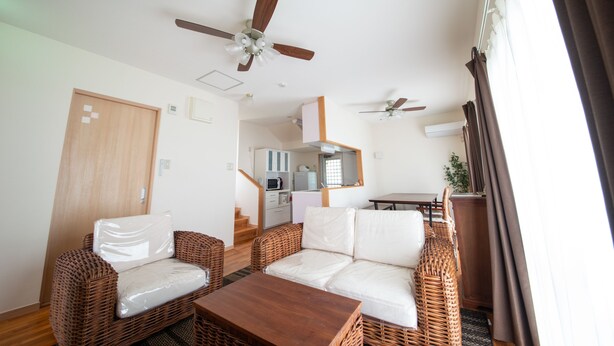 A room with wicker furniture, a wooden table, ceiling fans, and a kitchen area in the background. - Pine House in ISHIGAKI (Ishigaki)