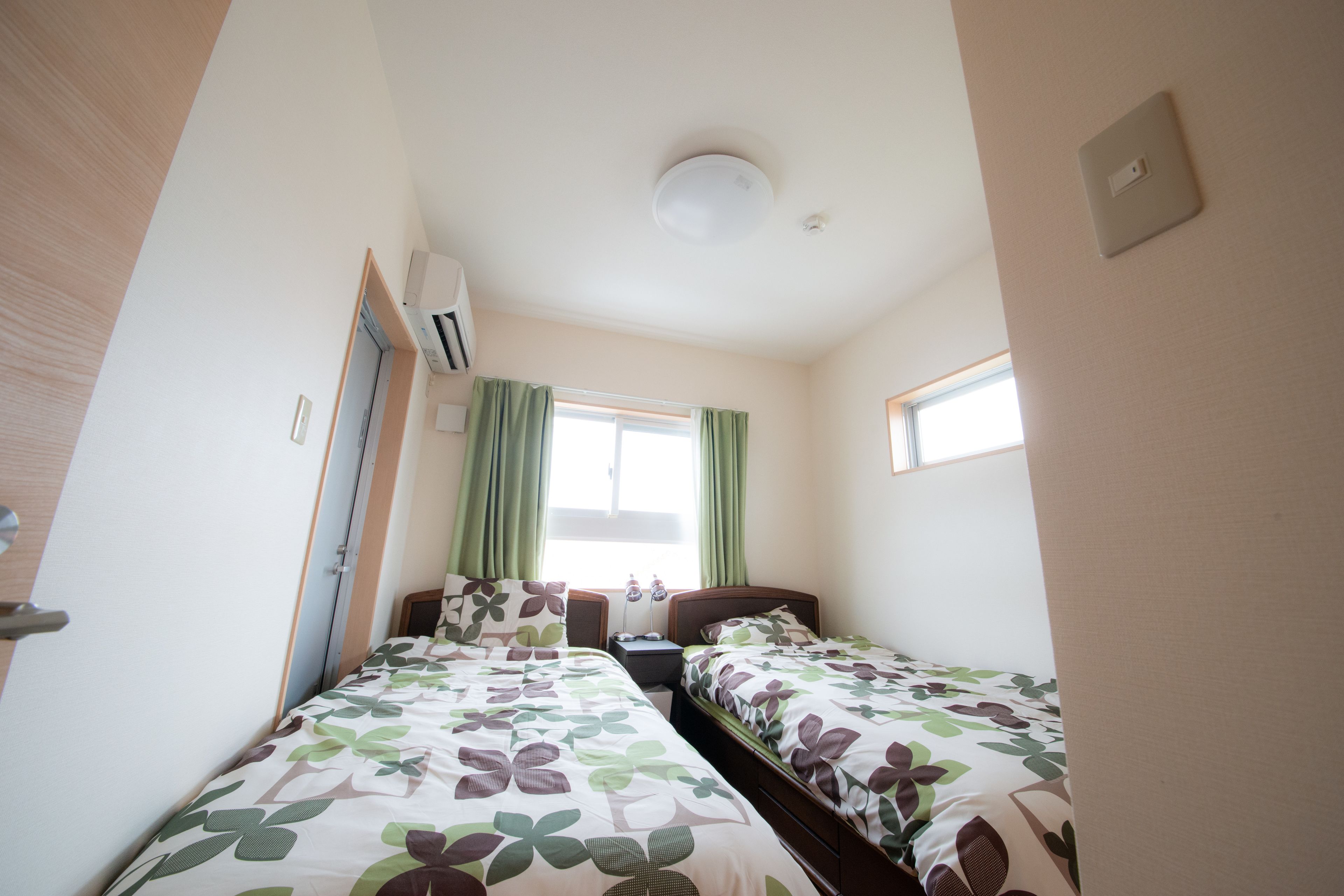A small, well-lit bedroom with two beds, a window with green curtains, and a ceiling light.