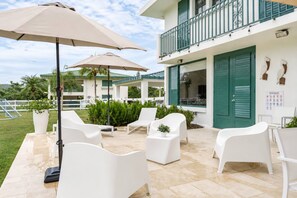 An outdoor seating area with white chairs and beige umbrellas, a pool, and a building with green shutters.