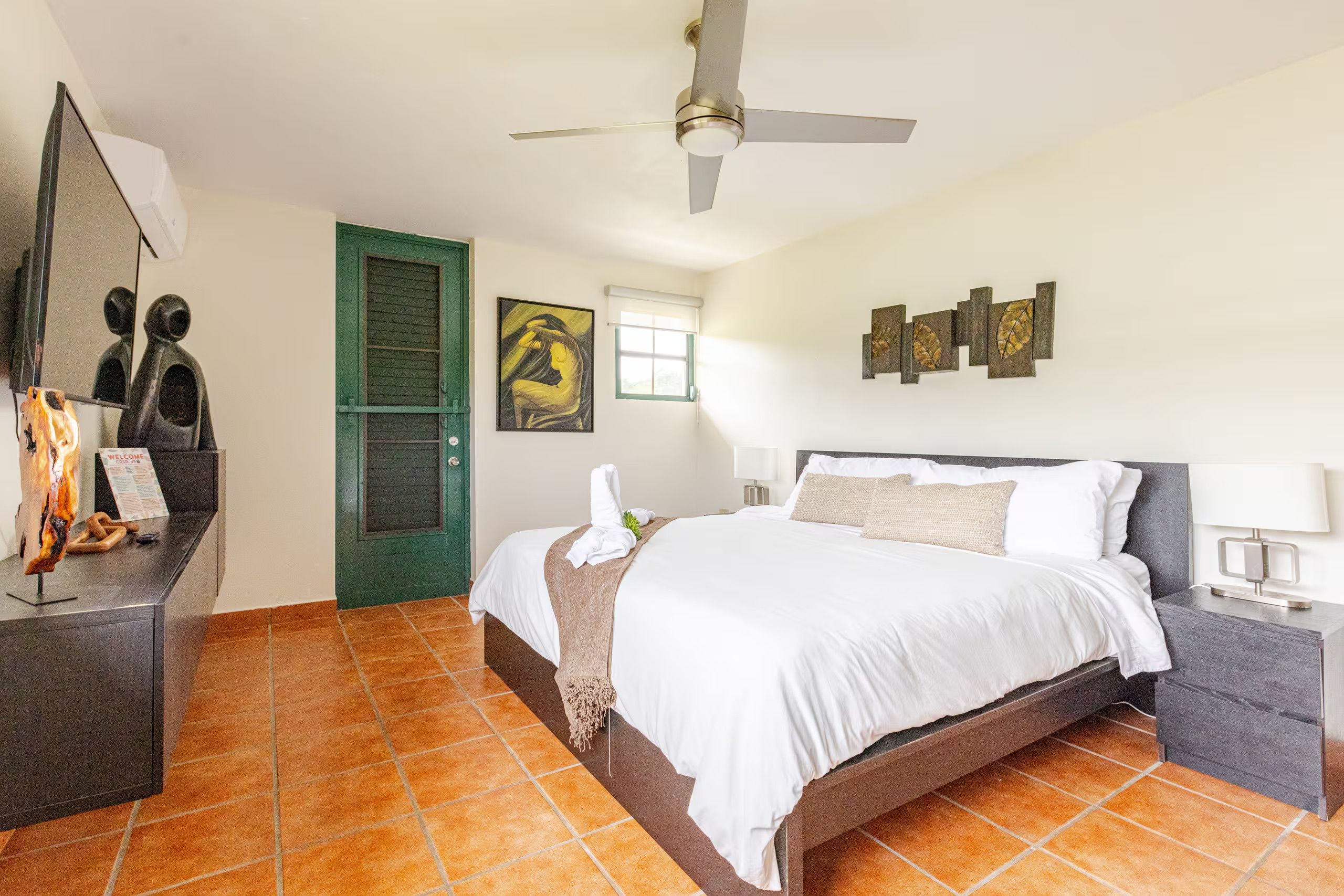 A hotel room with a bed, a desk, a green door, and artwork on the walls.