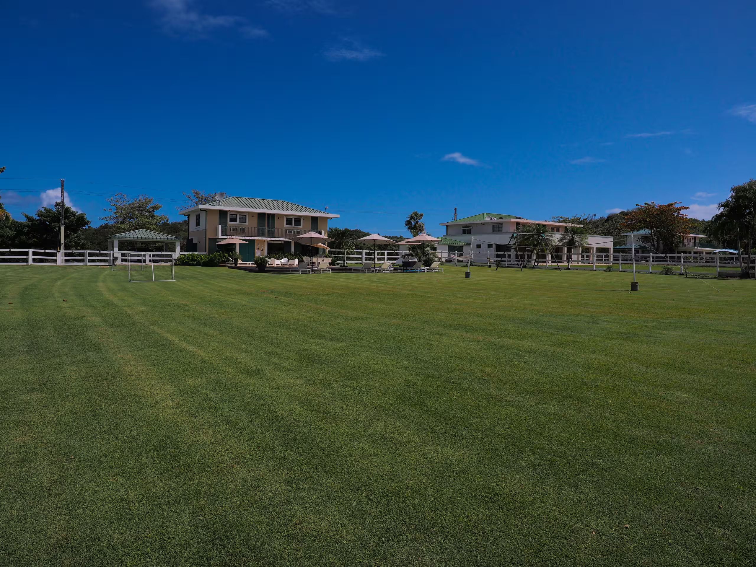 A well-maintained grassy field with a white fence, a gazebo, and a building with a green roof.