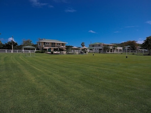 A well-maintained grassy field with a white fence, a gazebo, and two buildings with balconies and umbrellas.