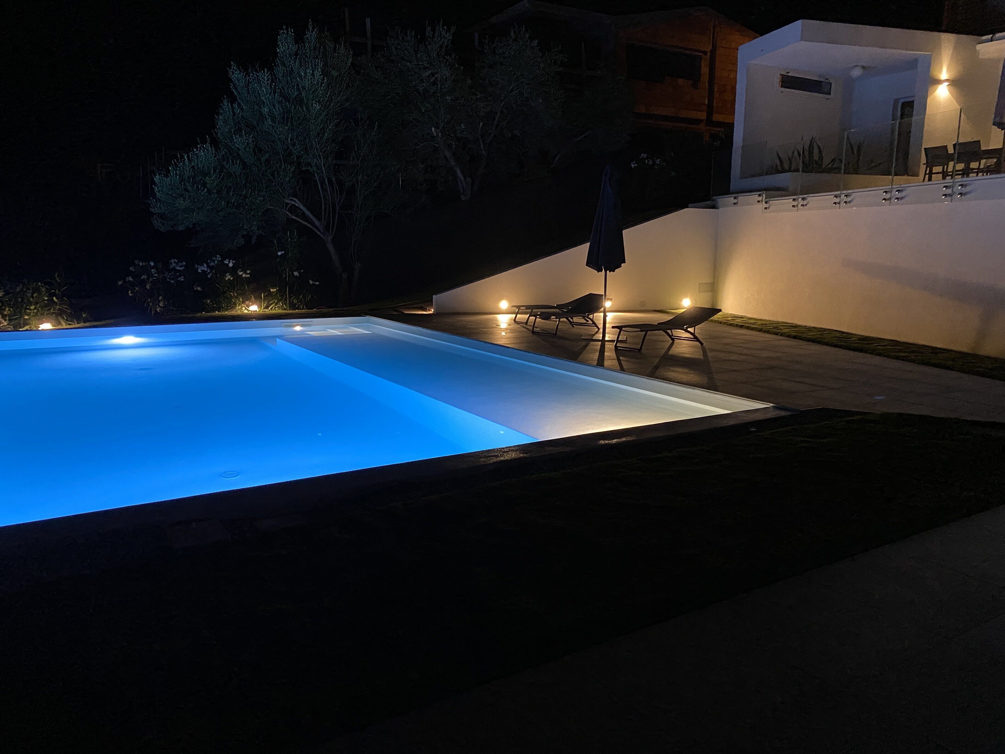 A swimming pool with a clear blue edge, surrounded by a patio area with lounge chairs and a wall-mounted umbrella.