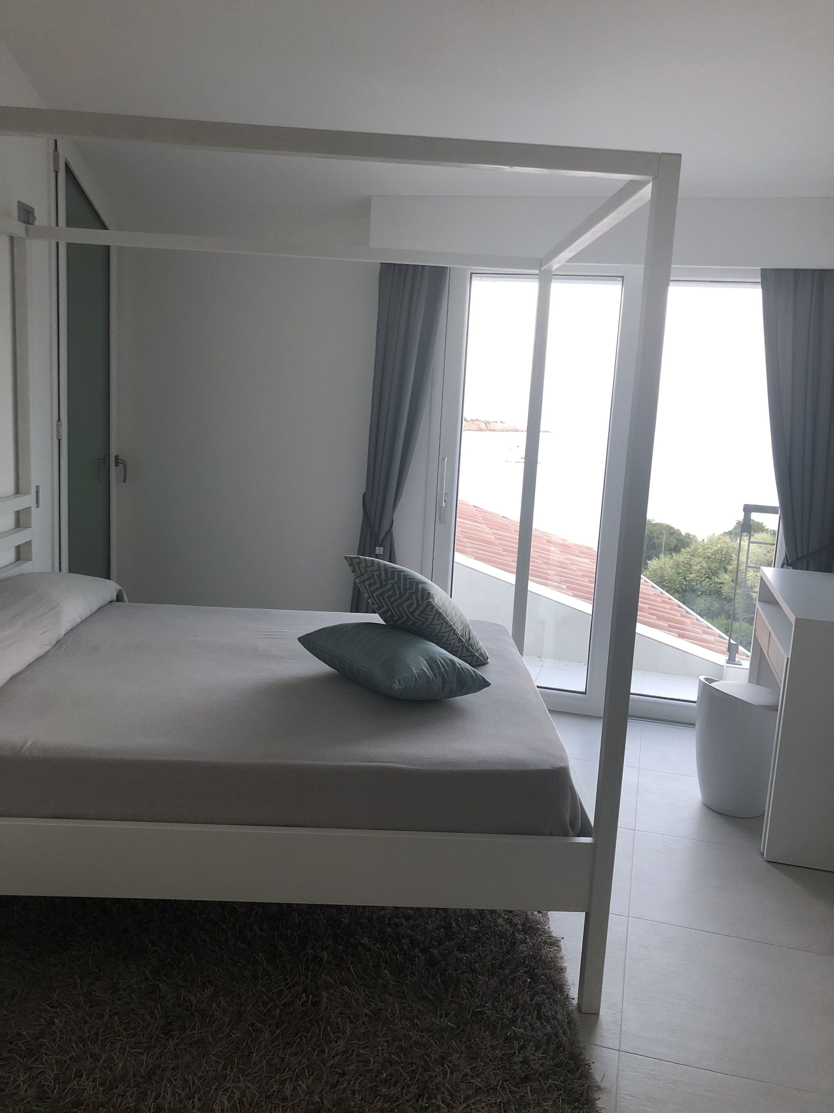 A modern bedroom with a bed, a small bedside table, a window with curtains, and a view of the outside.