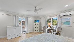 Une chambre avec un lit, un ventilateur de plafond, une télévision, un bureau avec des chaises et une fenêtre donnant sur l’extérieur.
