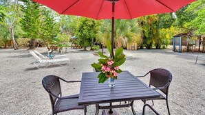 Une terrasse extérieure avec une table et des chaises sous un parasol rouge, entourée de verdure et recouverte de gravier.