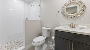 Une salle de bain avec des toilettes blanches, une douche, un meuble-lavabo avec des placards sombres et un miroir décoratif.