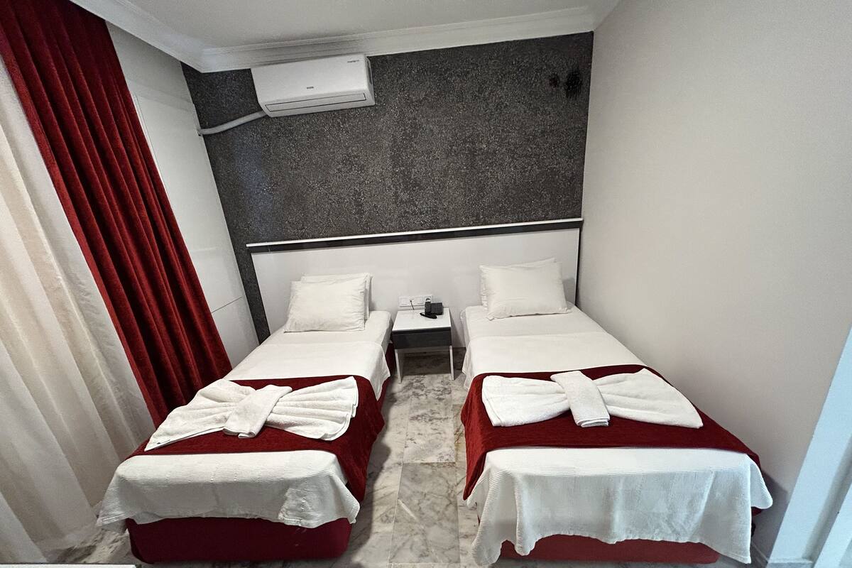 A hotel room with two beds, each with a white bedspread and a red blanket. There is a nightstand between the beds with a phone and a lamp. The room has a marble floor and a gray textured wall above the beds.