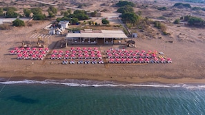 An aerial view of a beach with rows of sun loungers and umbrellas, a building with a flat roof, and a clear view of the ocean. - Bellissima Hotel (Manavgat)