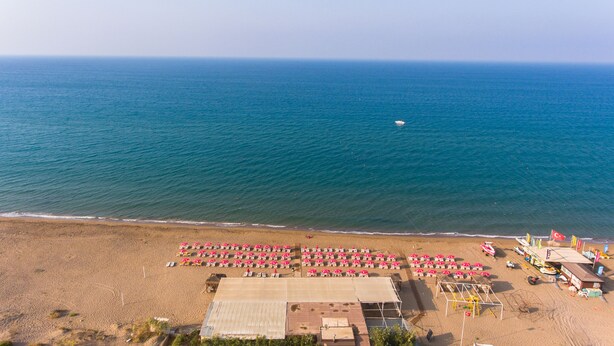An aerial view of a beach with rows of sun loungers and umbrellas, a small boat on the water, and a building with a roof. - Bellissima Hotel (Manavgat)