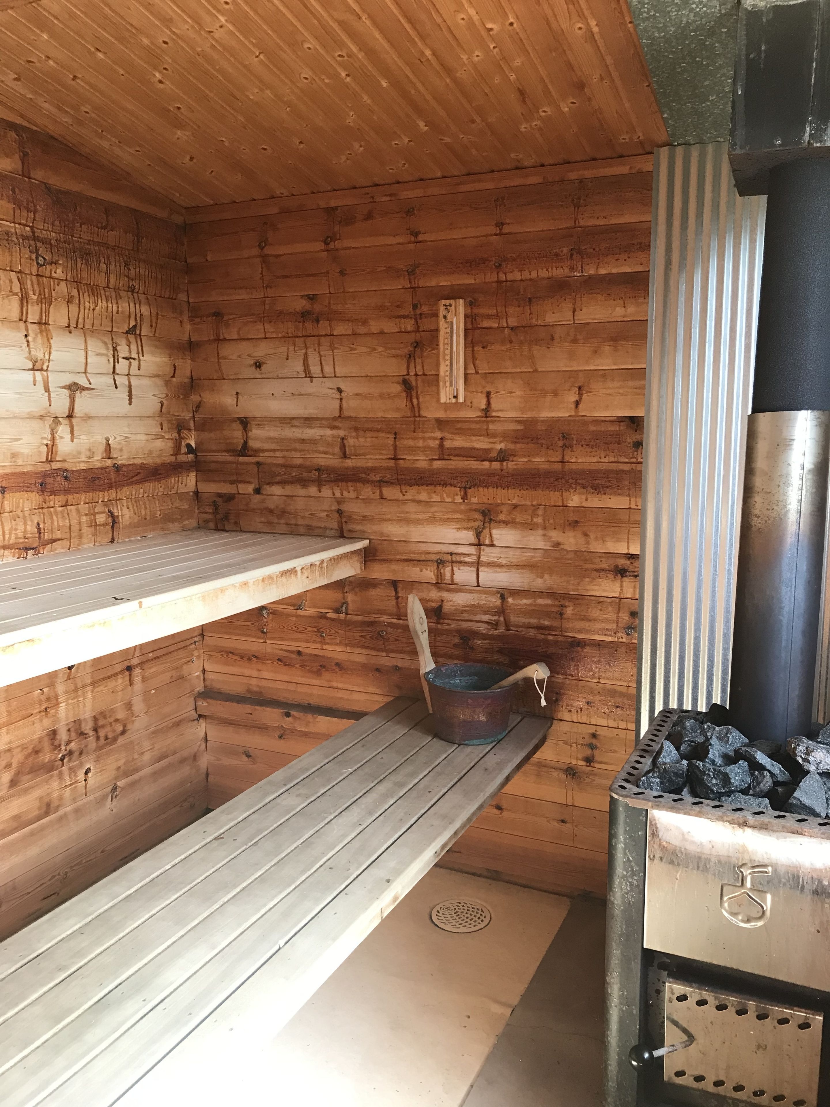 A wooden cabin interior with a metal stove, a wooden bench, and a metal bucket with a wooden spoon.