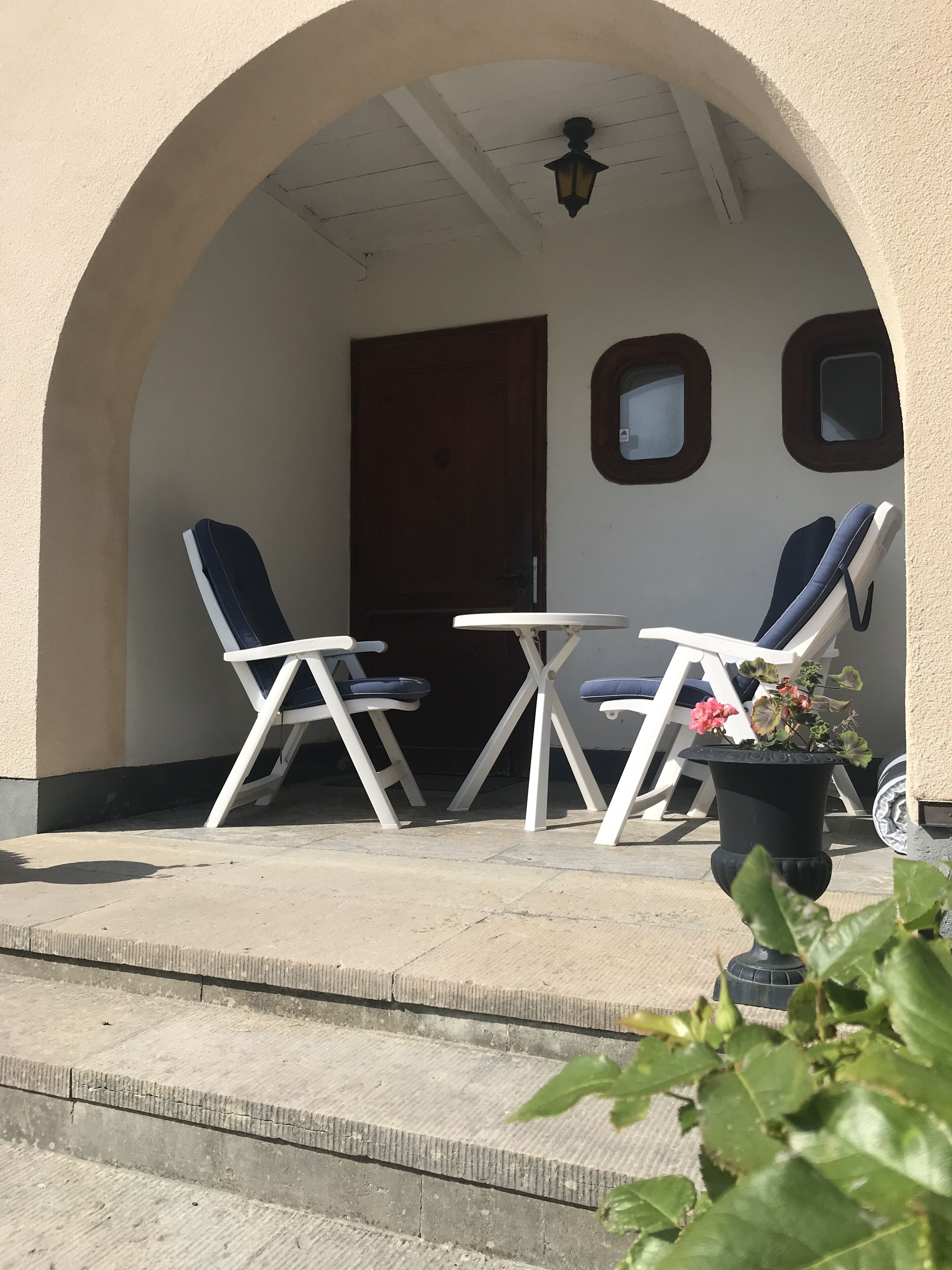 An outdoor seating area with two white tables and chairs, a potted plant, and a door.