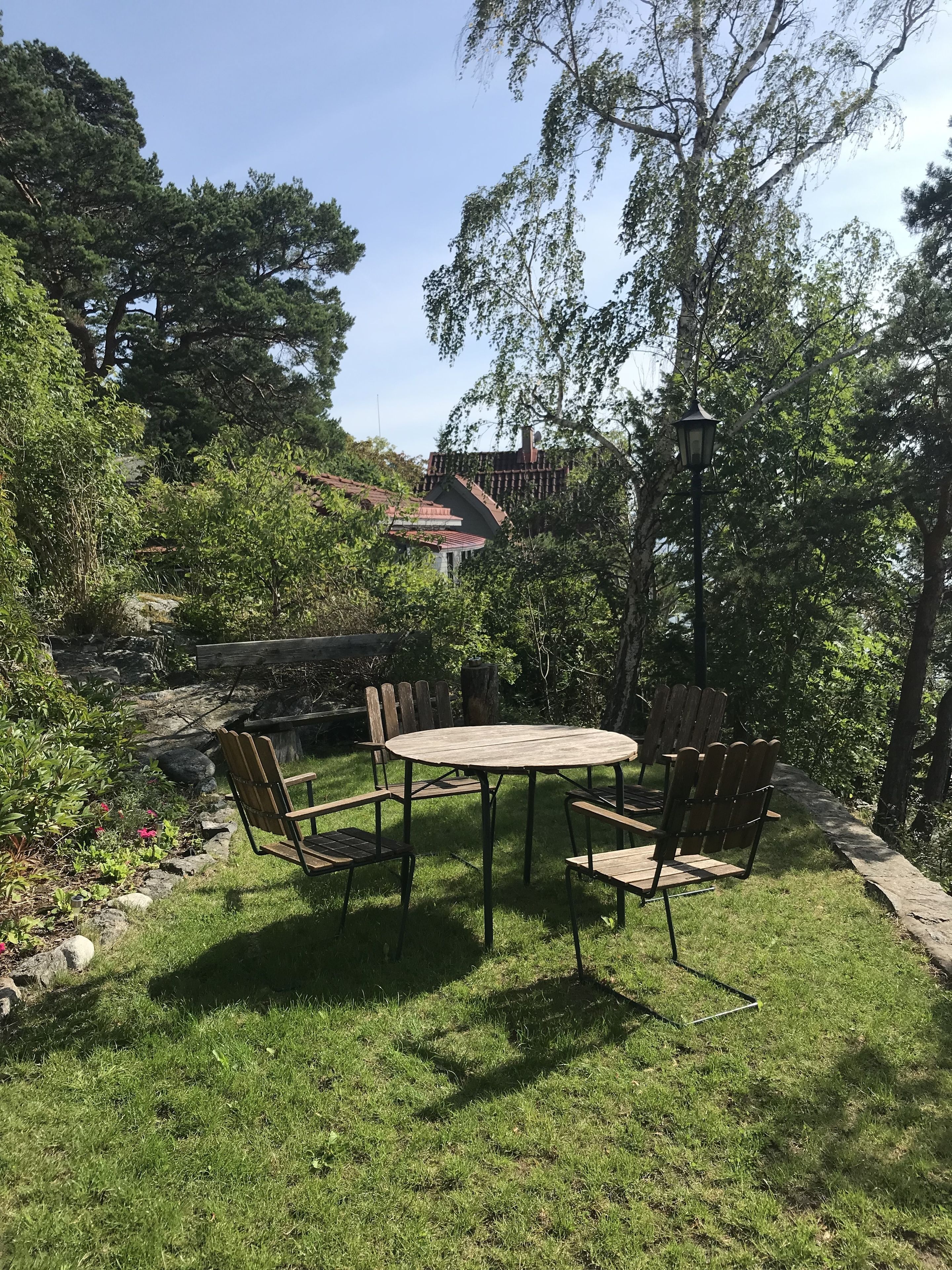 A round wooden table with metal chairs in a grassy outdoor area, surrounded by trees and a stone wall.