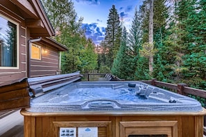 Un jacuzzi rodeado por una terraza de madera, con un paisaje boscoso y una cabaña.