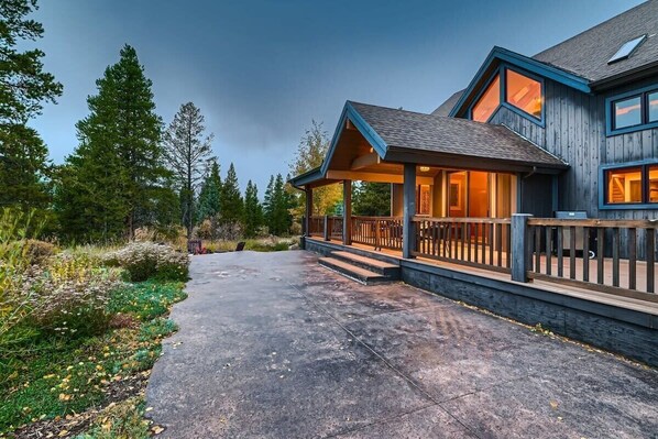 A two-story house with a covered porch, a concrete driveway, and a forested backdrop.