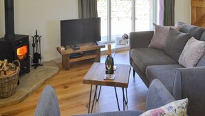 A cozy living room with a wood-burning stove, a television on a wooden stand, a sofa, and a table with a bottle and glass.