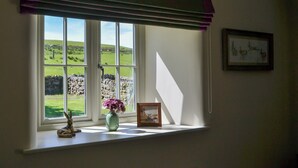 A window with a view of a green landscape, a stone wall, and a vase with flowers on the windowsill.