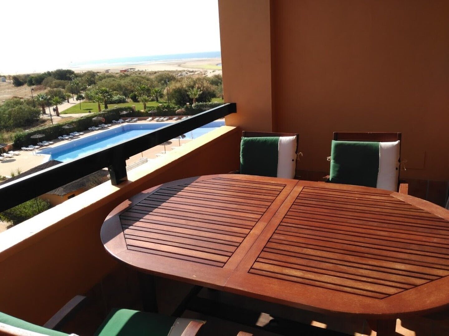 Een balkon met een houten tafel en twee stoelen, met uitzicht op een zwembad en een strand.