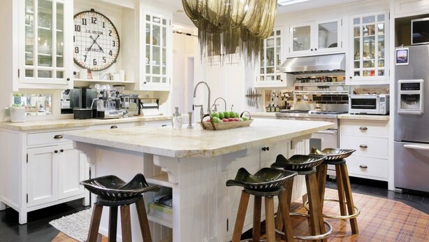 Une cuisine moderne avec des meubles blancs, un grand îlot central doté de tabourets, un lustre suspendu et une horloge au mur.
