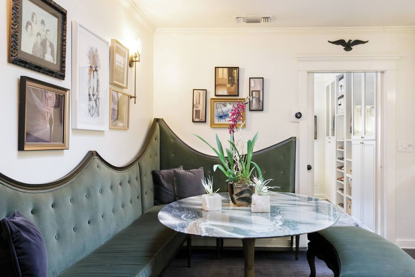 Una habitación con un banco tapizado en verde, una mesa redonda de vidrio con un florero y cuadros en la pared.