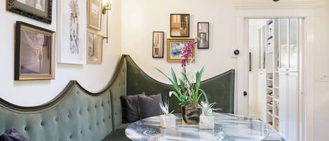Una habitación con un banco tapizado en verde, una mesa redonda de vidrio con un florero y cuadros en la pared.