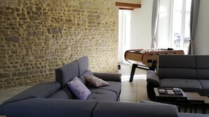 Ein modernes Wohnzimmer mit Steinwand, einem grauen Ecksofa, einem Kicker und einem großen Fenster mit Vorhängen.